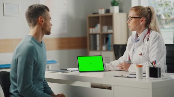 女医生在健康门诊向年轻男性病人展示绿色屏幕的平板电脑。坐在医院办公室桌子后面的家庭医生。