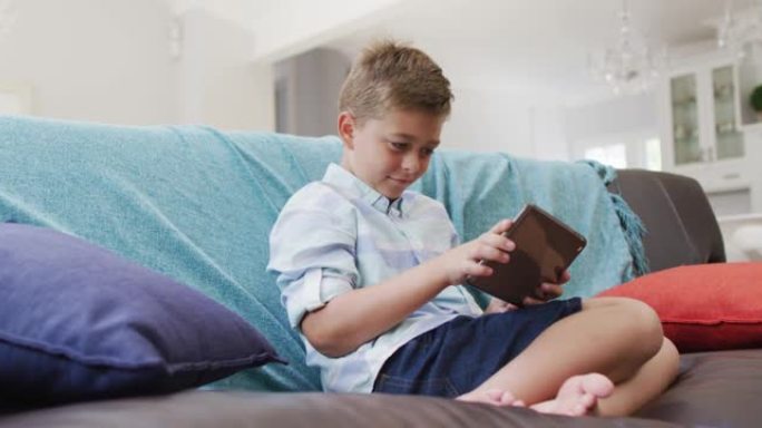 快乐的高加索男孩在家，坐在沙发上使用平板电脑，玩游戏并微笑