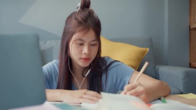 年轻的亚洲女孩少年，戴着休闲耳机，使用笔记本电脑在线学习在家里的客厅写讲座笔记本。隔离教育在线电子学
