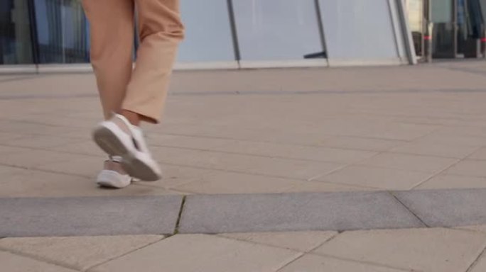 一名年轻女子穿着米色裤子和现代时尚的白色运动鞋走在广场上，从后面看