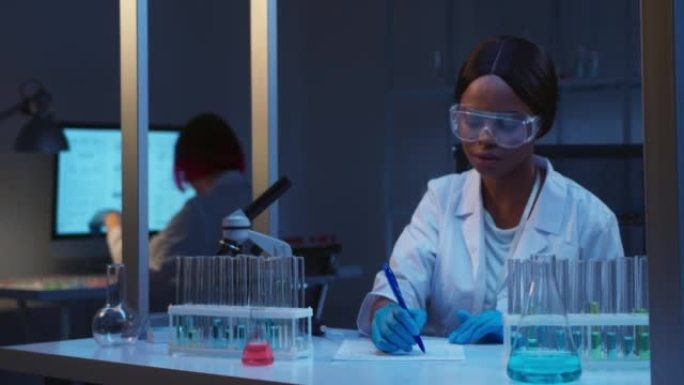 一位年轻的迷人的非裔美国女科学家戴着防护眼镜在现代实验室工作，她看着相机微笑