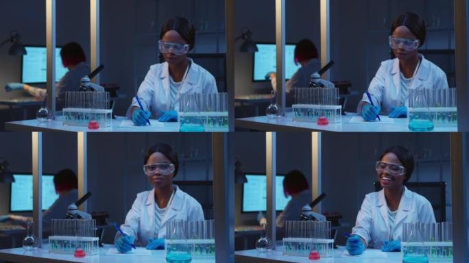一位年轻的迷人的非裔美国女科学家戴着防护眼镜在现代实验室工作，她看着相机微笑