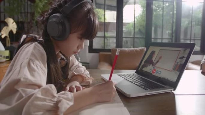 一个亚洲女孩正坐在家里专心地在线学习，并与哥哥在一起，通过平板电脑记下她所学到的知识。由于Covid