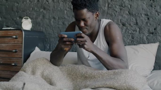 快乐的非洲裔美国人在家里床上用智能手机玩在线视频游戏的肖像