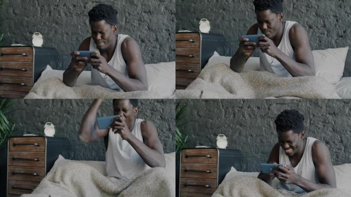 快乐的非洲裔美国人在家里床上用智能手机玩在线视频游戏的肖像