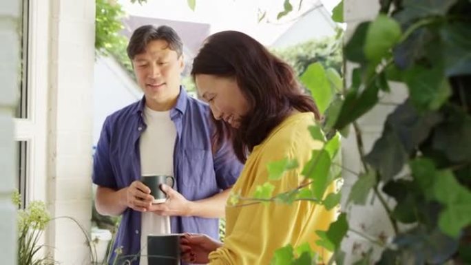 成熟的亚洲妇女在夏季花园里种植木制容器，丈夫在慢动作的杯子里喝热饮料