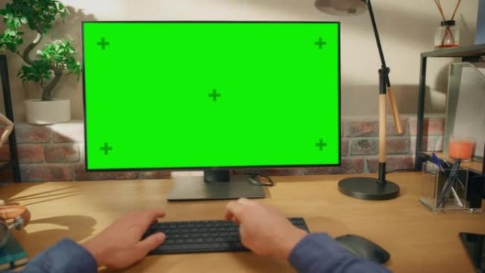 在具有绿屏模拟显示的台式计算机上工作的人的POV第一人称视图。男性坐在办公桌前，在时尚的客厅在家中打