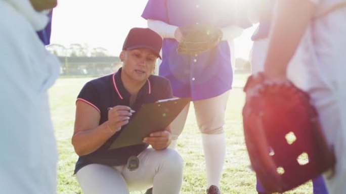 高加索女棒球教练蹲在剪贴板上写字，并在球场上与球队交谈