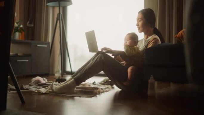 在家工作: 年轻的亚洲母亲的肖像，抱着可爱的婴儿，白天在笔记本电脑上工作。成功的女经理在远程工作的同