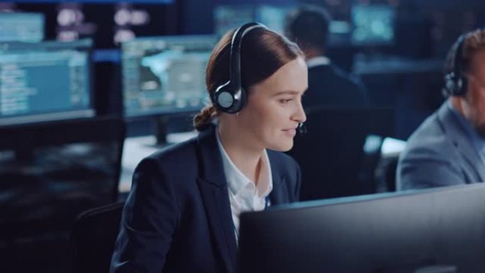 欢乐美丽的技术客户支持专家在一个充满同事和显示屏的黑暗监视和控制室中的计算机上工作时，正在耳机上聊天