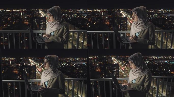 戴头巾的女人晚上在阳台上的笔记本电脑上打字