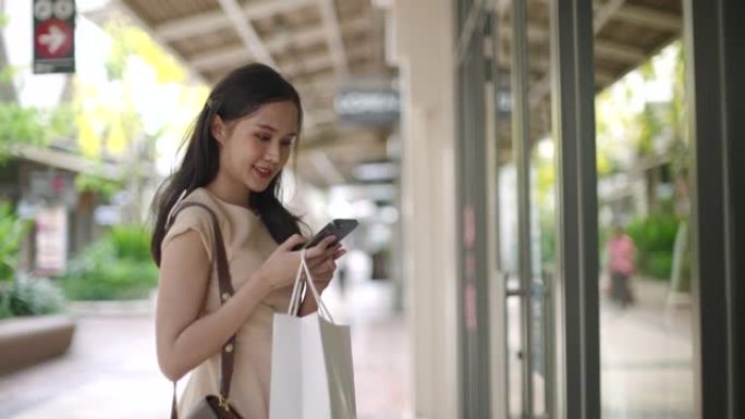 亚洲快乐女性使用带购物袋的智能手机看着商店橱窗