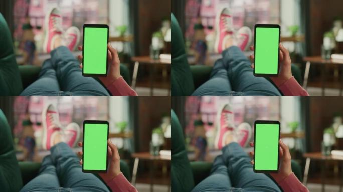 女性手握着带有绿屏模拟显示的智能手机。女性正在家里的沙发上放松，在移动设备上观看视频和阅读社交媒体帖