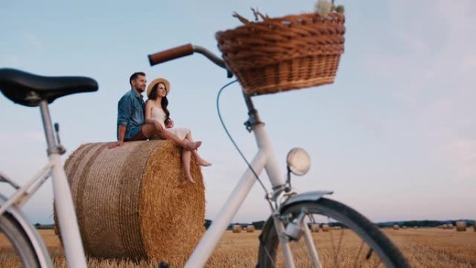 中年夫妇坐在干草捆上的手持慢动作镜头，自行车在前景中，欣赏日落