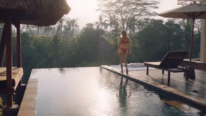 美丽的女人穿着比基尼在游泳池边放松，在豪华酒店水疗中心享受早晨日出，可以看到热带丛林
