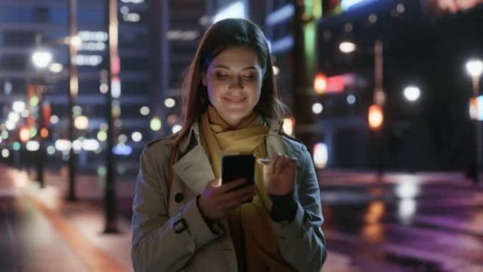 一个穿着风衣的美女的肖像在夜晚霓虹灯的现代城市街道上行走。迷人的女性使用智能手机，环顾城市电影环境。