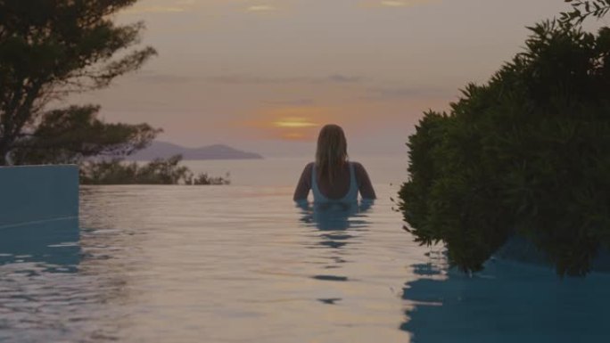 日落时在无边泳池中享受游泳的女人的SLO MO肖像