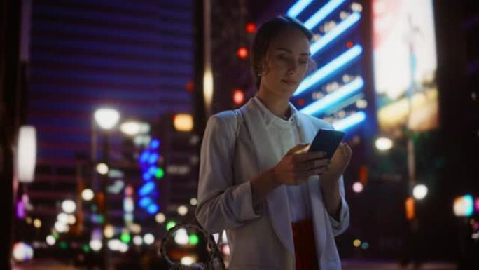 美丽的年轻女子使用智能手机走过充满霓虹灯的夜晚城市街道。微笑着若有所思的女性使用手机，发布社交媒体，