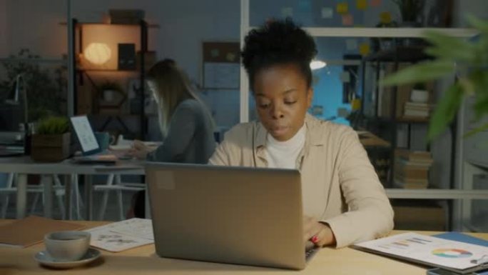 疲倦的非裔美国女商人使用笔记本电脑，晚上在黑暗的办公室里为巨大的工作量感到压力