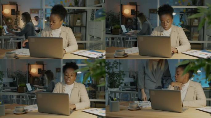 疲倦的非裔美国女商人使用笔记本电脑，晚上在黑暗的办公室里为巨大的工作量感到压力