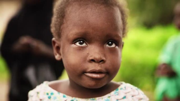 一个可爱的非洲小托德勒对着镜头微笑的特写肖像，背景模糊的人在移动。代表对美好未来的希望的黑人农村儿童