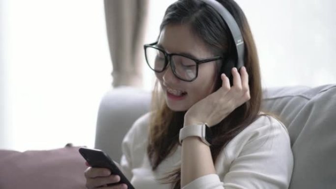 年轻的亚洲女性在客厅放松，用智能数字设备上的耳机听音乐