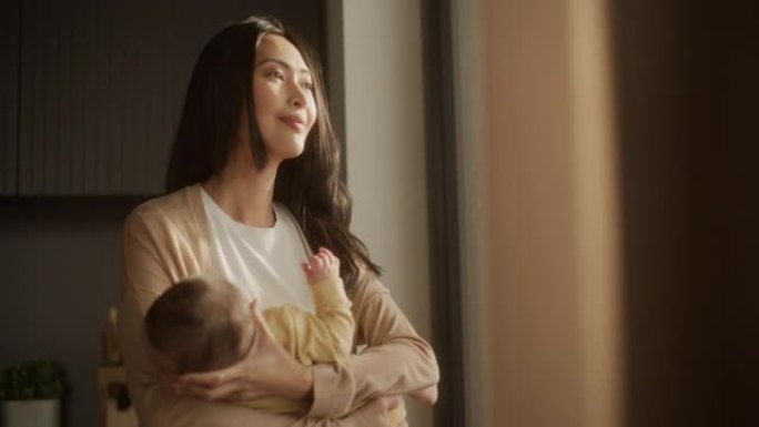 一位亚洲母亲的肖像将婴儿抱在怀里，透过家里的一扇大窗户看着街道。平静的女人喜欢看雨，和蹒跚学步的孩子
