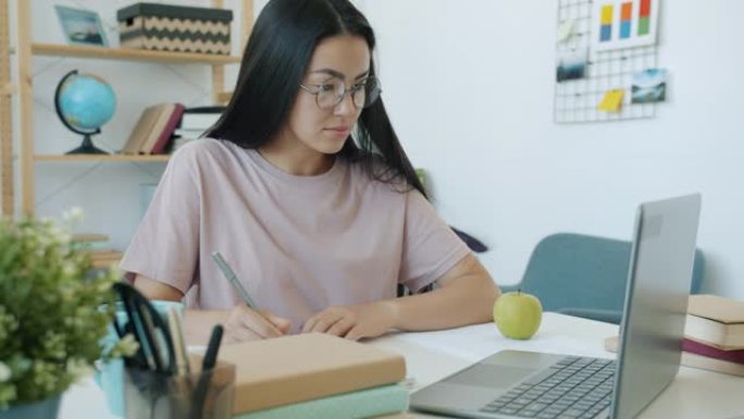 美丽的亚洲女孩在家学习写作，看着桌子上的笔记本电脑屏幕