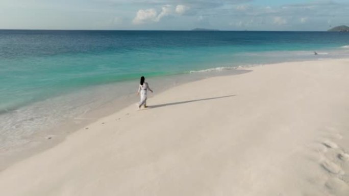 一名妇女在热带岛屿上的碧绿海水中沿着原始海滩的水边行走的空中后视