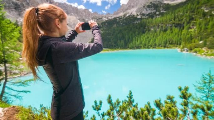在多洛米蒂山湖索拉皮斯的年轻女徒步旅行者在晴天用智能手机拍照