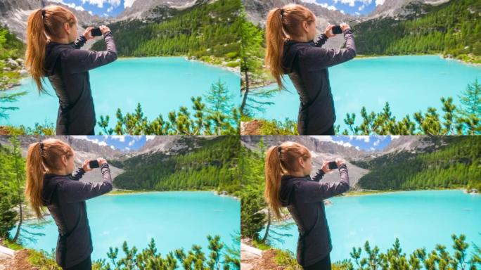 在多洛米蒂山湖索拉皮斯的年轻女徒步旅行者在晴天用智能手机拍照