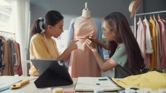 两位年轻的亚洲女士裁缝看着数字平板电脑用针将织物附着在人体模特上，创造服装设计新系列在商店分享创意。