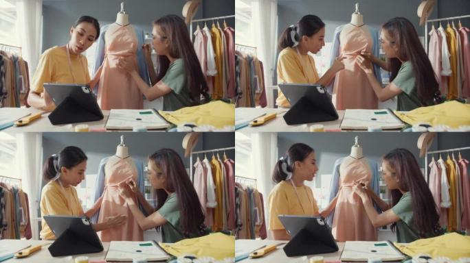 两位年轻的亚洲女士裁缝看着数字平板电脑用针将织物附着在人体模特上，创造服装设计新系列在商店分享创意。