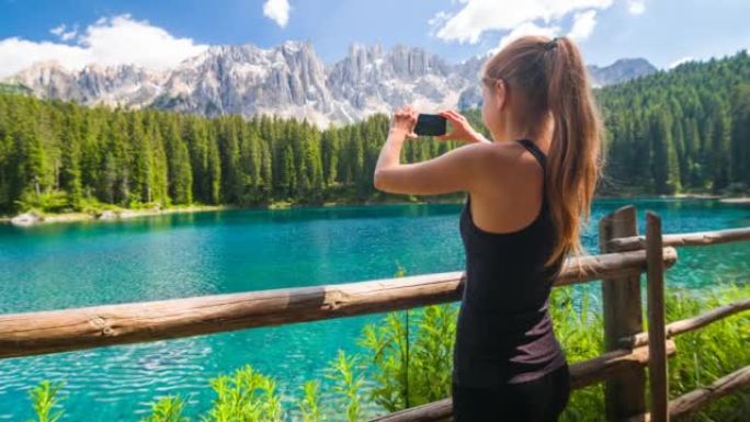 年轻的女性游客探索多洛米蒂山腰，站在特伦蒂诺的卡雷扎湖旁，背景是拉特马尔山脉，用智能手机拍照
