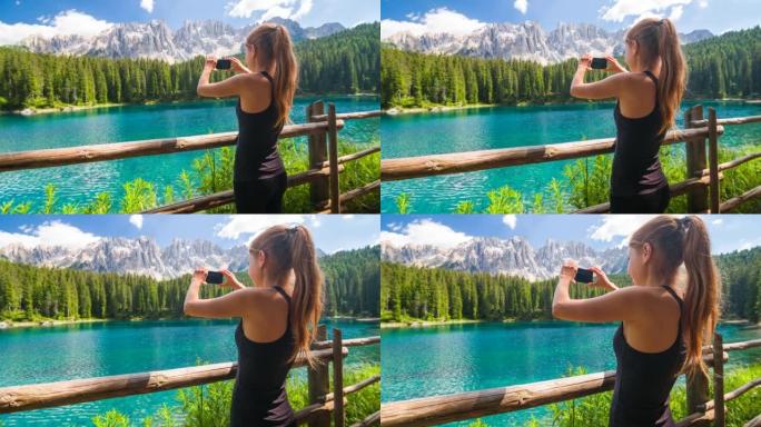 年轻的女性游客探索多洛米蒂山腰，站在特伦蒂诺的卡雷扎湖旁，背景是拉特马尔山脉，用智能手机拍照