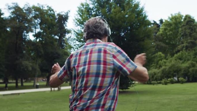 一个活跃的老人在公园里活泼跳舞的肖像，用耳机听音乐。快乐开朗的老年男性，穿着一件彩色衬衫享受生活，伴