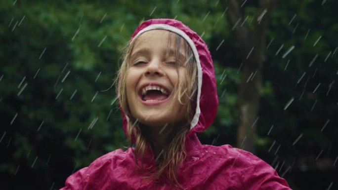 一个穿着粉红色雨衣的快乐小女孩在雨中快乐地站着，被大自然和树木包围着。女孩微笑着，在雨天独自在外面玩