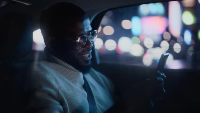 戴着眼镜的快乐黑人晚上坐出租车后座回家。英俊的男性使用智能手机，在城市街道上开着霓虹灯的汽车时微笑。