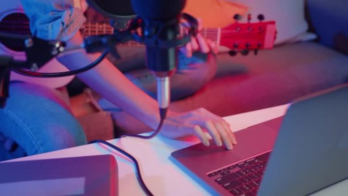 快乐亚洲女孩博主弹吉他，晚上在现代客厅家庭工作室的笔记本电脑上使用麦克风唱歌录音音乐混音器。