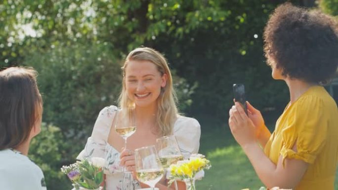女人在手机上拍照的朋友坐在夏天的花园里喝着酒，欢呼着庆祝，同时聊天和慢镜头拍摄