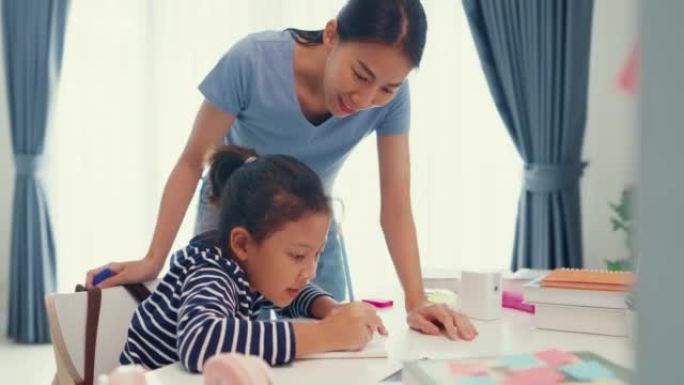 穿着毛衣的亚洲蹒跚学步的女孩坐在桌子前，拿着记事本，用铅笔专注于写笔记本，周末在家和妈妈或家教做数学