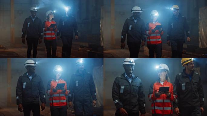 三名多元文化的重工业工程师和穿着制服的工人在黑钢工厂里用手电筒戴着安全帽行走。女性工业承包商正在使用