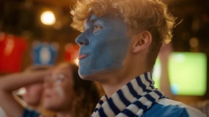 特写一位年轻英俊的足球迷的肖像，脸上涂着蓝色和白色的脸，站在酒吧里的人群中，高呼，跳跃，为足球队欢呼