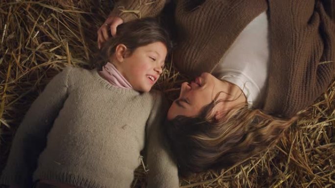 电影拍摄的快乐微笑的母亲和小女儿躺在农村农田谷仓的干草上，一起开心地说话和大笑。爱情、农业、家庭、母