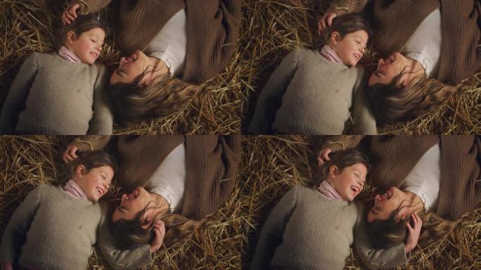 电影拍摄的快乐微笑的母亲和小女儿躺在农村农田谷仓的干草上，一起开心地说话和大笑。爱情、农业、家庭、母