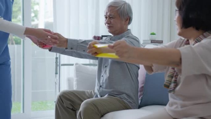 年轻的老年亚洲公民夫妇坐在沙发沙发上配合居家护理，辅助生活，搓洗护士使用阻力带练习为老年患者在家中进