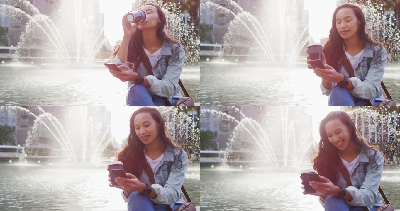 亚洲妇女坐在喷泉旁，使用智能手机喝外卖咖啡