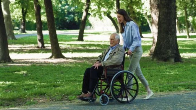 无忧无虑的快乐孙女和坐在轮椅上的祖父在绿色公园一起散步的电影镜头。幸福，自由，生活，祖父母，世代，爱