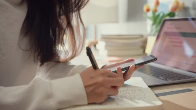 特写年轻的亚洲女士自由职业聚焦手机聊天同事工作财务在家里的客厅笔记本上写作。女学生在家在线学习，在家
