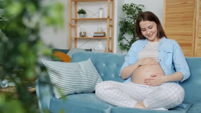 怀孕的年轻女子向婴儿表达爱意，抚摸着肚子微笑着坐在家里的沙发上